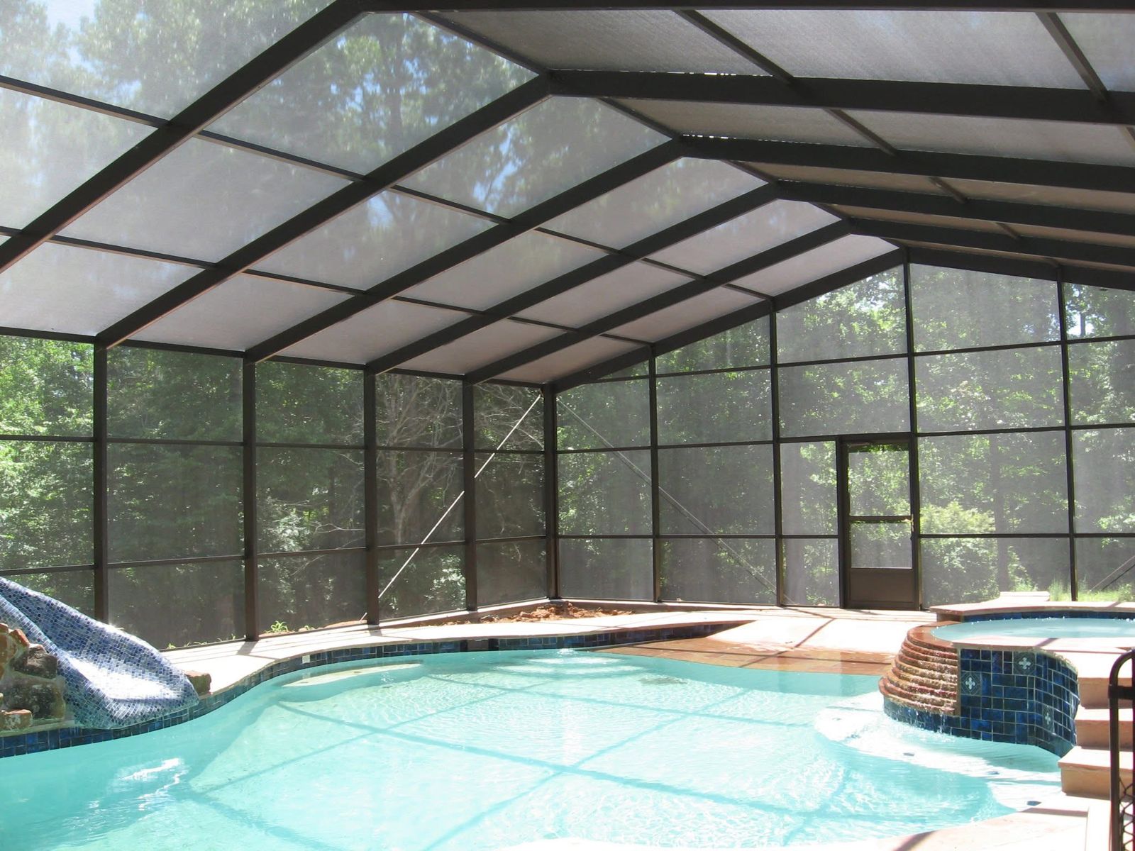 Inside a dark bronze pool screen enclosure with a gabled roof frame over a pool and spa