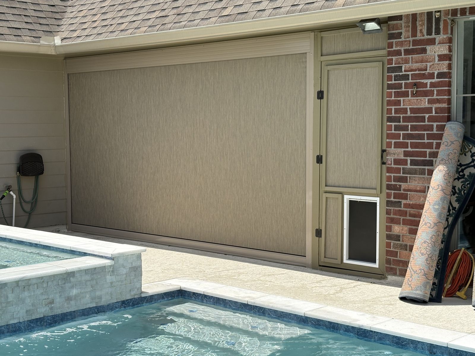 Tan motorized shade beside a screen door with a pet door next to a pool